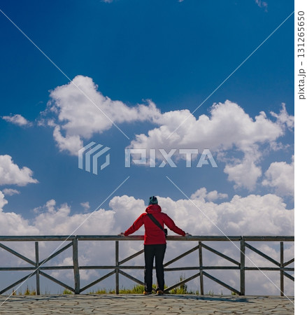 Individual stands on a balcony in a red jacket, gazing towards a partially cloudy sky, embodying a sense of contemplation. Wooden railing frames the expansive view. Individual stands on a balcony in a red jacket, gazing towards a partially cloudy sky, embodying a sense of contemplation. Wooden railing frames the expansive view. 131265650