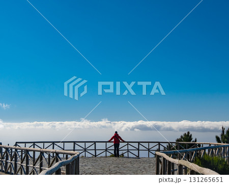 Individual stands on a balcony in a red jacket, gazing towards a partially cloudy sky, embodying a sense of contemplation. Wooden railing frames the expansive view. 131265651