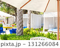 Luxury cabanas or tent with white curtains and blue lounge chairs under palm trees and umbrellas. Green plants and inflatable unicorn float create tropical summer resort atmosphere. Summer, beach, 131266084