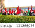 UAE flags waving in the wind in front of a modern glass building with geometric patterns, green hedge and palm trees under bright sunlight. Holiday, state event 131266086
