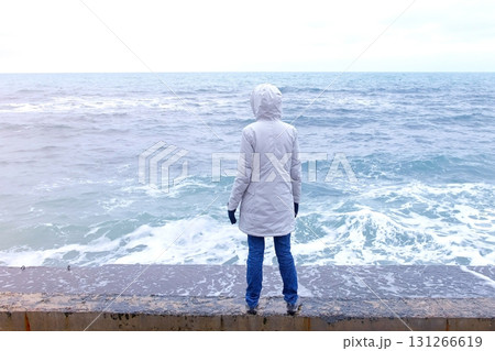Woman in a white jacket stands on the waterfront on the beach looks at storm waves. Back view. 131266619