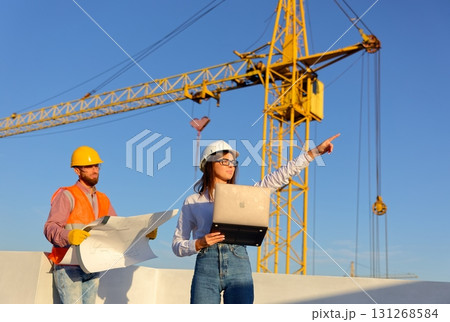 Engineers discussing blueprint on construction site with crane and laptop Engineers discussing blueprint on construction site with crane and laptop 131268584