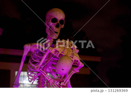 A tall Halloween skeleton holds a small baby skeleton while purple light glows on the lawn. Spooky outdoor decoration creates a dramatic, eerie yard scene. 131269369