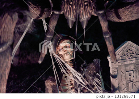 A Halloween skeleton sits beneath a giant spider with fangs, webs, and red eyes beside an RIP tombstone. Spooky yard decoration with dark lighting on a suburban lawn. A Halloween skeleton sits beneath a giant spider with fangs, webs, and red eyes beside an RIP tombstone. Spooky yard decoration with dark lighting on a suburban lawn. 131269371