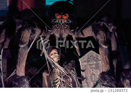 A huge hairy spider with red eyes looms over a skeleton beside an RIP tombstone. Creepy Halloween yard decoration with webs, fangs, and eerie lighting on the lawn. 131269372