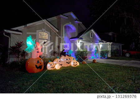 A suburban house displays glowing inflatables with pumpkins and ghosts at night. Skull light projections, spider webs, and a black cat create a festive, spooky holiday scene. A suburban house displays glowing inflatables with pumpkins and ghosts at night. Skull light projections, spider webs, and a black cat create a festive, spooky holiday scene. 131269376
