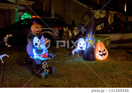 Blue alien skeleton, ghost, pumpkin, and spooky tree inflatables glow on a suburban lawn at night. Halloween yard decorations with bright lights create a festive, eerie holiday display. 131269388