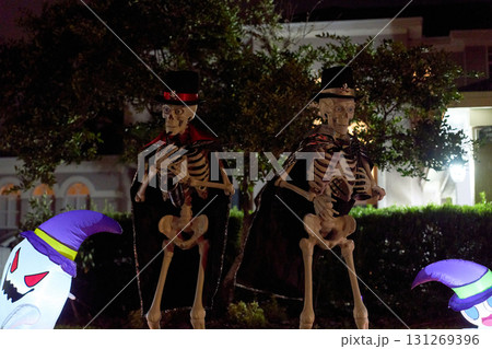 Two skeletons in top hats and capes pose with instruments on the grass. Spooky night yard display with glowing inflatables nearby. Two skeletons in top hats and capes pose with instruments on the grass. Spooky night yard display with glowing inflatables nearby. 131269396