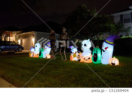 A row of glowing inflatable ghosts and pumpkins lines a suburban lawn with two skeleton figures. Festive neighborhood Halloween display after dark. 131269404