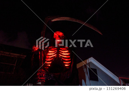 Hooded skeleton reaper glows red with bright ribs and eyes beside a house. Dramatic Halloween yard display with large scythe and deep shadows. Hooded skeleton reaper glows red with bright ribs and eyes beside a house. Dramatic Halloween yard display with large scythe and deep shadows. 131269406