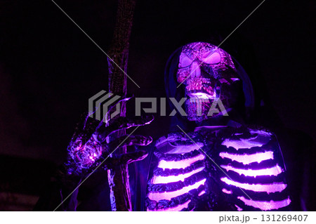 Hooded skeleton reaper holds a staff with purple neon light at night. Glowing ribs and claws create a spooky Halloween yard display. Hooded skeleton reaper holds a staff with purple neon light at night. Glowing ribs and claws create a spooky Halloween yard display. 131269407