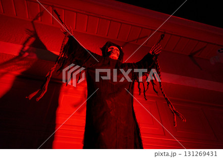 A hooded skeleton in tattered robes hangs with arms raised under strong red light. Spooky Halloween yard decoration with dramatic shadows at night. 131269431