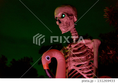 A large skeleton figure with glowing eyes stands at night holding a pink flamingo prop. Festive Halloween yard display with dramatic green sky and colorful lighting. A large skeleton figure with glowing eyes stands at night holding a pink flamingo prop. Festive Halloween yard display with dramatic green sky and colorful lighting. 131269433