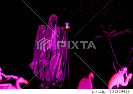 Two tall hooded reapers with lanterns glow purple in a spooky lawn scene. The nighttime haunted house yard is filled with eerie lights and shadows. Two tall hooded reapers with lanterns glow purple in a spooky lawn scene. The nighttime haunted house yard is filled with eerie lights and shadows. 131269438
