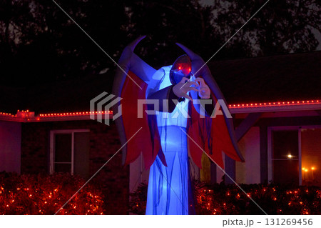 A towering blue demon with red eyes and wide wings stands glowing in a decorated yard. The giant inflatable creates a scary Halloween display at night. A towering blue demon with red eyes and wide wings stands glowing in a decorated yard. The giant inflatable creates a scary Halloween display at night. 131269456
