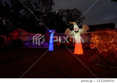Front yard house displays a glowing inflatable ghost and a tall blue winged demon at night. Colorful lights and bushes create a spooky Halloween display. 131269459