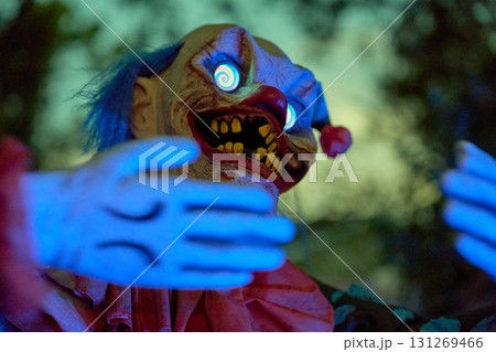Creepy clown reaches forward with blue-lit hands and hypnotic spiral eyes. Red nose, yellow teeth, and dramatic lighting create a scary Halloween scene. Creepy clown reaches forward with blue-lit hands and hypnotic spiral eyes. Red nose, yellow teeth, and dramatic lighting create a scary Halloween scene. 131269466