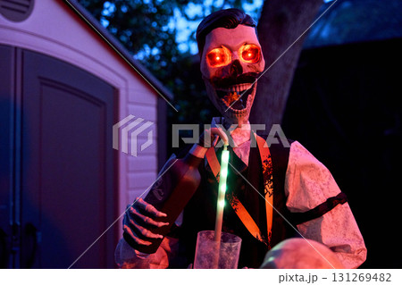 A skeleton bartender pours a drink into a glass at night. Red glowing eyes and neon lighting create a spooky party scene. 131269482