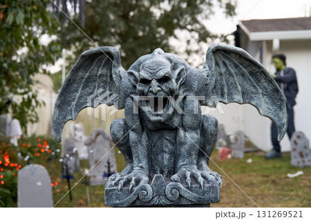 A winged stone gargoyle crouches on a pedestal in a Halloween graveyard. Tombstones, lights, and a spooky yard create a haunted outdoor scene. 131269521