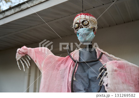 Grim skeleton figure in tattered pink robe wears a stained face mask and head wrap. Spooky hospital-inspired Halloween decoration displayed outside a home. Grim skeleton figure in tattered pink robe wears a stained face mask and head wrap. Spooky hospital-inspired Halloween decoration displayed outside a home. 131269582