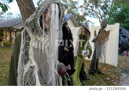 Hooded ghoul stands in the foreground while several witches gather behind a white fence near a house. Outdoor Halloween yard display with orange lights and spooky props. 131269584