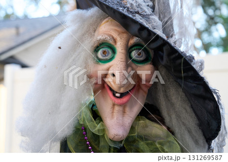 Grinning witch prop with wild white hair, green eye makeup, and a tall black hat. Spooky Halloween yard decoration photographed outdoors in daylight. 131269587
