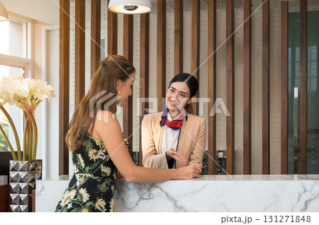 Cheerful hotel receptionist in uniform engages female guest in friendly conversation at marble front desk for hotel check in 131271848