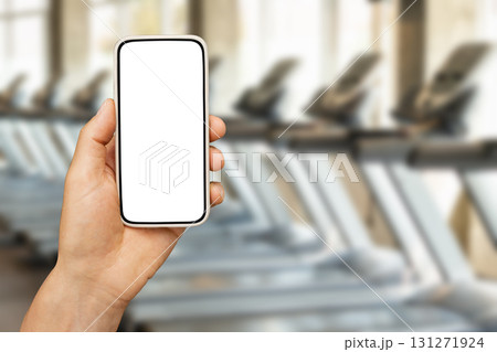 Mans hand holding smartphone with white blank screen against treadmill at gym. Mockup for design. 131271924
