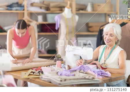Senior woman sewing create wardrobe items, female assistant drawing paper dress pattern by curves Senior woman sewing create wardrobe items, female assistant drawing paper dress pattern by curves 131275140