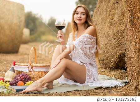 Sexy girl in transparent dress drinks red wine on picnic near hay bale Sexy girl in transparent dress drinks red wine on picnic near hay bale 131275365