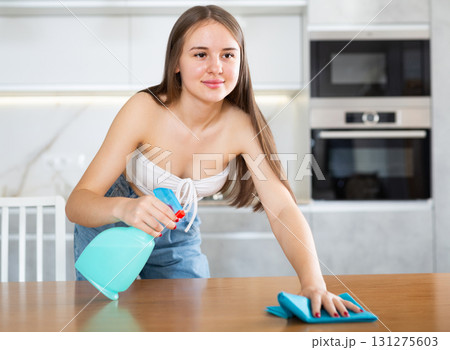 In kitchen, positive housewife in casual clothes holds rag in hands and cleans surface of table 131275603
