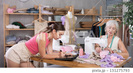 Elderly seamstress sews female assistant draws pattern on tablet 131275793