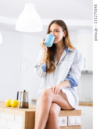 Portrait of woman sitting on table in kitchen and drinking coffee. Woman in shirt drinking coffee in morning Portrait of woman sitting on table in kitchen and drinking coffee. Woman in shirt drinking coffee in morning 131275848