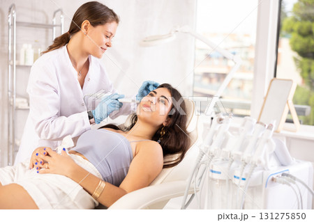 European aesthetician making facial beauty procedure with leather knife attachment to brunette girl 131275850