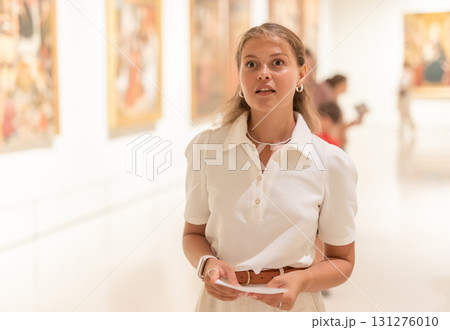 Young girl holding an information booklet looks at paintings in an art exhibition in museum Young girl holding an information booklet looks at paintings in an art exhibition in museum 131276010
