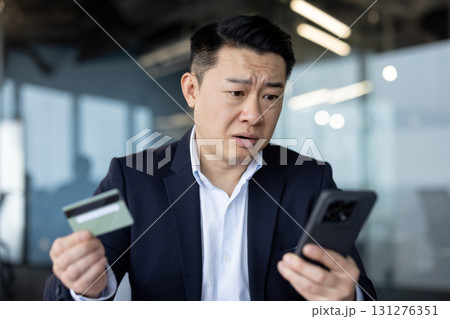 Upset young Asian man in a suit is in the office, holding a credit card and looking frustrated at the mobile phone screen. Upset young Asian man in a suit is in the office, holding a credit card and looking frustrated at the mobile phone screen. 131276351