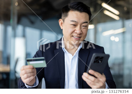 Close-up photo of a young Asian man in a suit sitting in an office, holding a credit card and using a mobile phone. 131276354