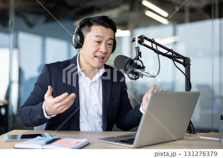 Asian young man in a business suit sitting in the office wearing headphones, in front of a microphone and talking on video call on a laptop. 131276379