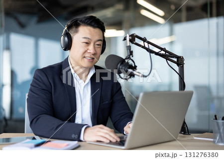 Smiling young Asian man wearing headphones sitting at a desk in an office in front of a microphone stand and working on a laptop. 131276380