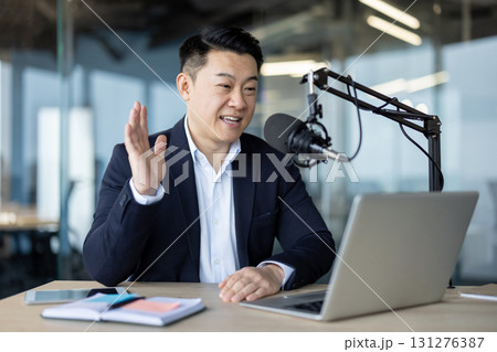 Asian young man in a suit sitting in the office in front of a desk with a microphone and talking, on a video call on a laptop. 131276387