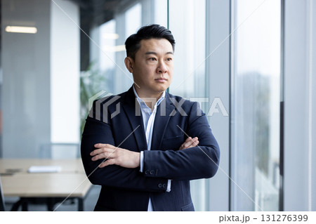 Asian young successful and serious businessman standing in a suit in the office, arms crossed on his chest, looking out the window. 131276399