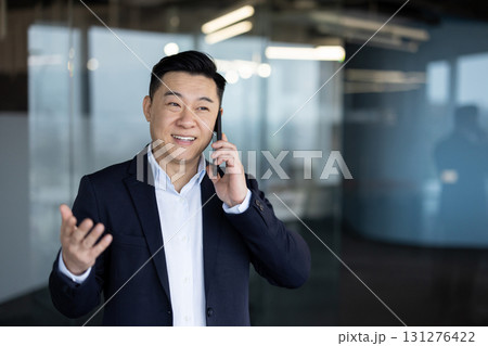 Young successful smiling Asian businessman in suit is in office and talking on mobile phone gesturing with hands. Young successful smiling Asian businessman in suit is in office and talking on mobile phone gesturing with hands. 131276422