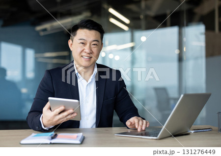 Portrait of a young Asian businessman sitting at a desk in the office, working on a laptop and using a tablet. Portrait of a young Asian businessman sitting at a desk in the office, working on a laptop and using a tablet. 131276449