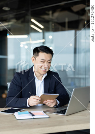 Vertical photo of an Asian young man working in an office at a laptop, smiling at the screen and taking notes in a notebook. 131276486