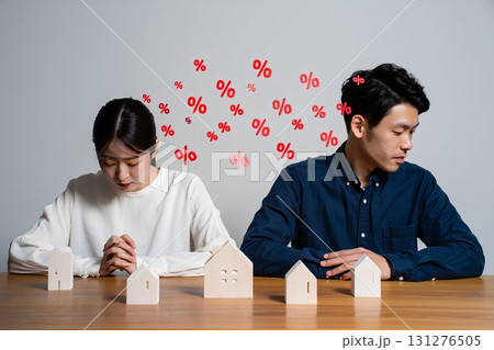 Stressed young couple looking at miniature house models with floating interest rate percentages, burden of high mortgage interest rates 131276505