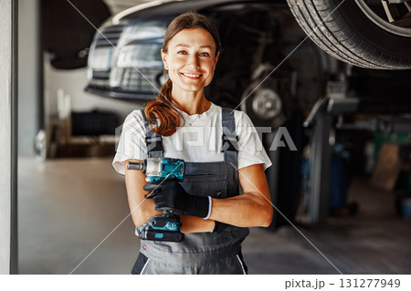 A smiling female mechanic holds a power tool in an auto repair shop, showcasing her skills A smiling female mechanic holds a power tool in an auto repair shop, showcasing her skills 131277949