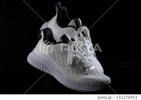 pair of white sneakers featuring a textured upper and black accents rests on a dark surface. The laces are untied adding a casual look. The sneakers highlight contemporary athletic footwear design. 131278451