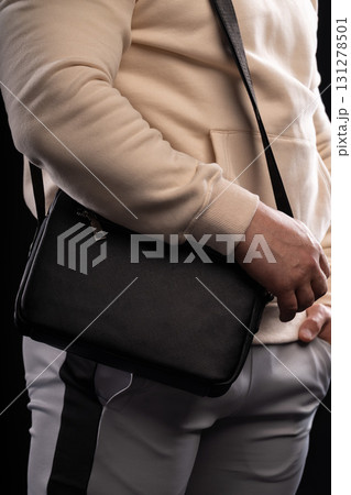 man in a beige hoodie stands with a black bag slung over his shoulder in a studio. His casual style contrasts with a sleek background creating a modern look. man in a beige hoodie stands with a black bag slung over his shoulder in a studio. His casual style contrasts with a sleek background creating a modern look. 131278501