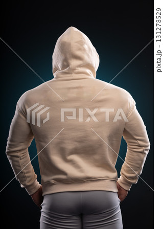 athletic man stands with his back facing the camera wearing a beige hoodie. The backdrop is dark highlighting his toned figure and the casual style of the outfit. 131278529