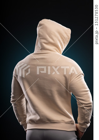 athletic man is facing away wearing a beige hoodie with hands on his hips. The dark background highlights his strong physique and creates a dramatic effect in the studio. 131278530